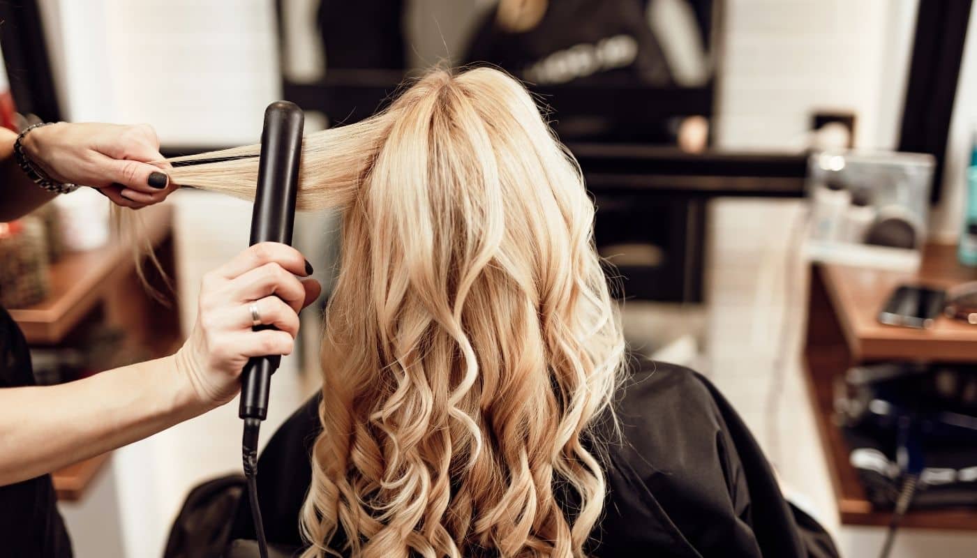 Stylist using a flat iron to curl a client's blonde hair in a hair salon