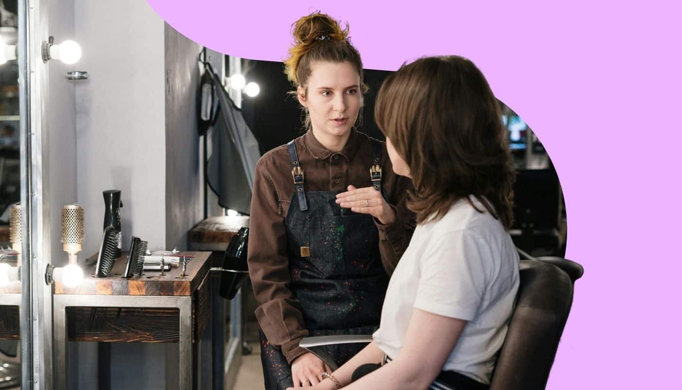 Hairstylist consults with a client seated in a salon chair, discussing hair goals and service options before an appointment. The image features a lavender branded background and a professional salon setting with illuminated mirrors, styling tools, and workstations, emphasizing personalized client consultations and salon expertise.
