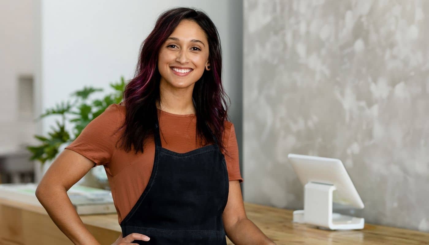 Smiling salon professional stands behind a reception counter wearing a black apron, posing in a modern salon. A point of sale system, wood countertop, and contemporary interior design elements are visible in the background.