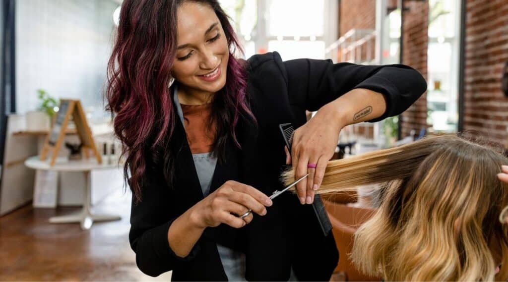 A smiling hairstylist with dark, wavy hair and purple highlights trims the blonde ends of a client’s hair inside a bright, modern salon.
