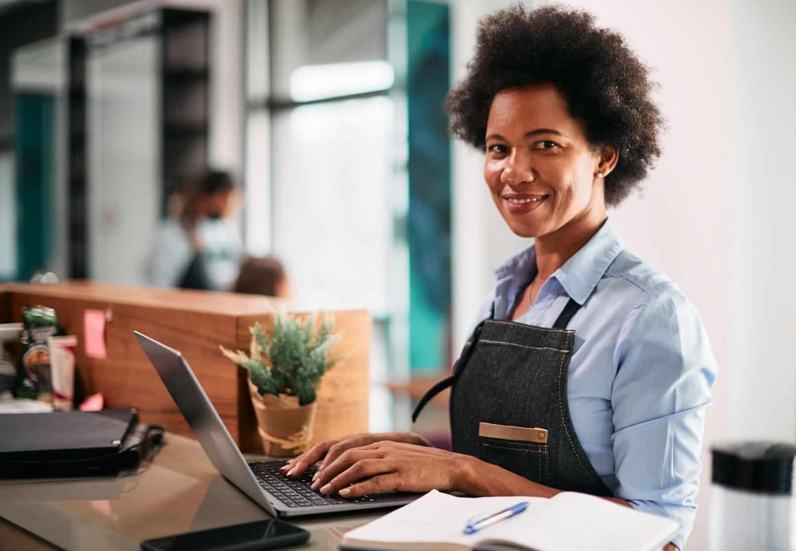 A salon professional wearing an apron works on a laptop at the front desk, surrounded by styling tools and a clean, contemporary workspace