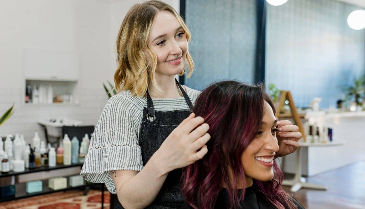 Hairstylist styles a client’s freshly colored hair while both smile in a bright, modern salon. Hair care products, plants, and salon stations are visible in the background, creating a professional and welcoming atmosphere.