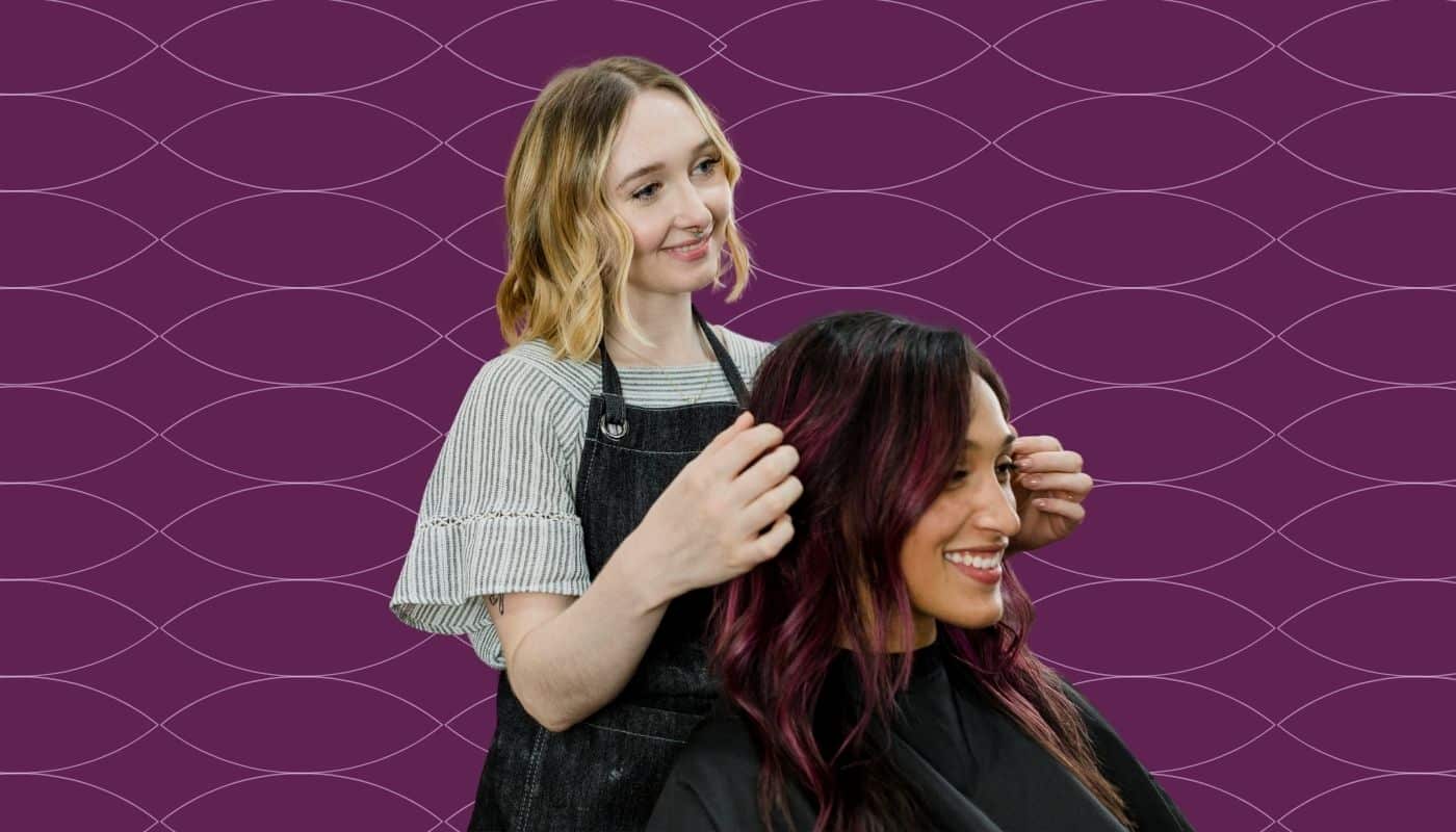 Hairstylist styles a client’s vibrant burgundy hair while both smile, set against a purple branded background with a subtle wave pattern. The image highlights personalized salon services, client care, and professional hairstyling expertise.