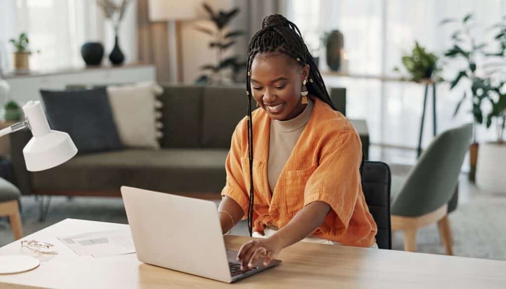 Professional working on a laptop at a desk in a modern home office. The workspace features a desk lamp, documents, indoor plants, and comfortable seating in the background, creating a productive and organized environment.