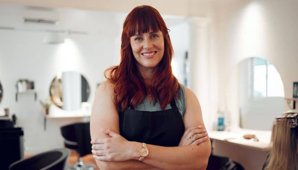 Smiling salon professional with red hair stands with arms crossed in a bright, modern salon. Styling stations, mirrors, and salon chairs are visible in the background, creating a welcoming and professional atmosphere.