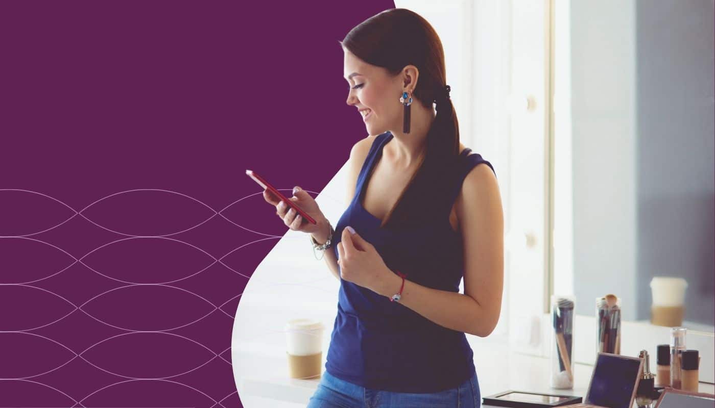Woman stands in a salon space checking her smartphone while smiling. Beauty products, makeup brushes, and a coffee cup sit on a nearby counter, while a purple branded graphic with a subtle wave pattern frames the image, highlighting salon technology, client communication, and business growth tools.