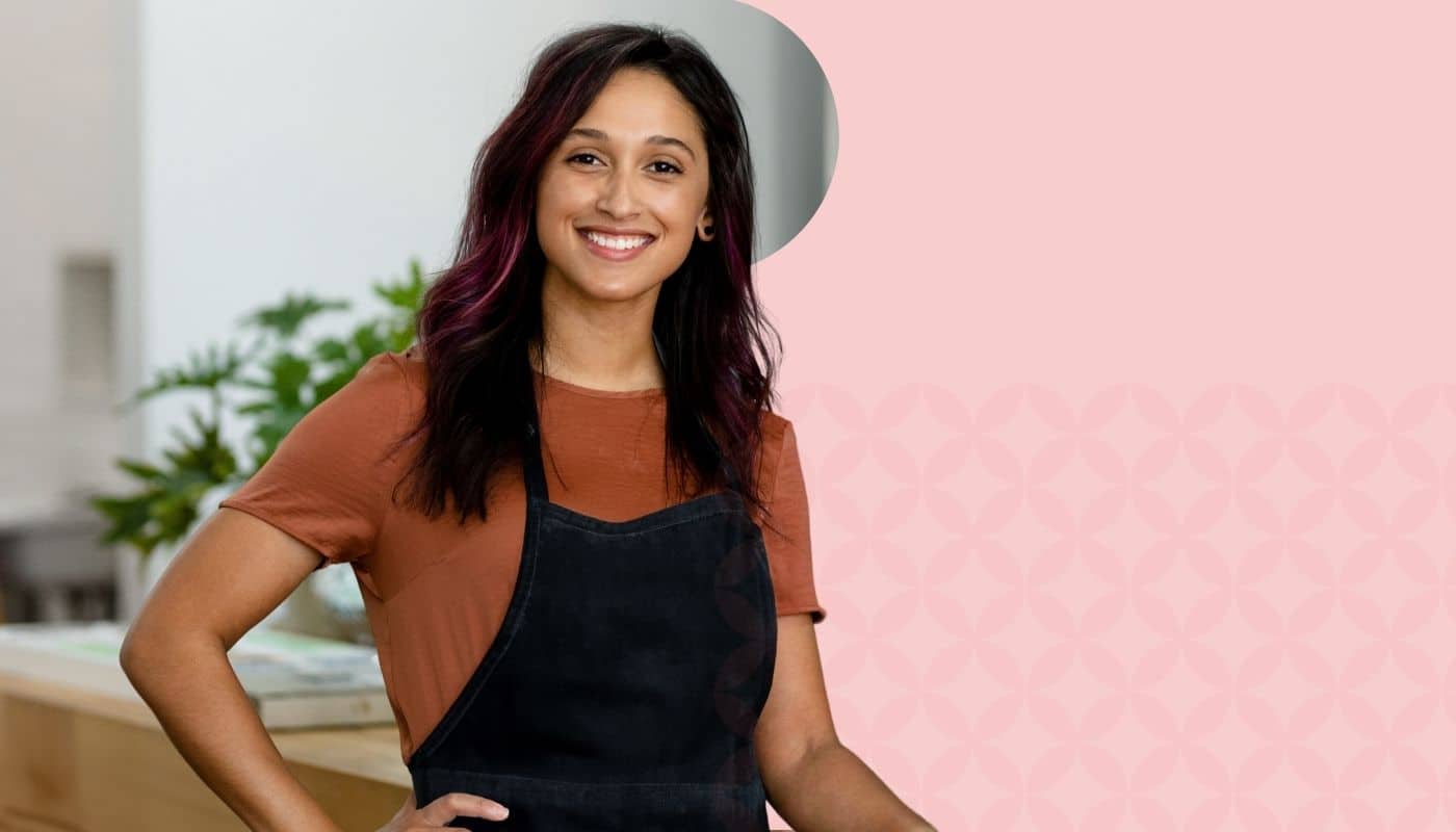 Smiling salon professional stands confidently in a modern salon wearing a black apron. The image features a soft pink branded background with subtle geometric patterns, highlighting salon business management, professionalism, and beauty industry expertise.