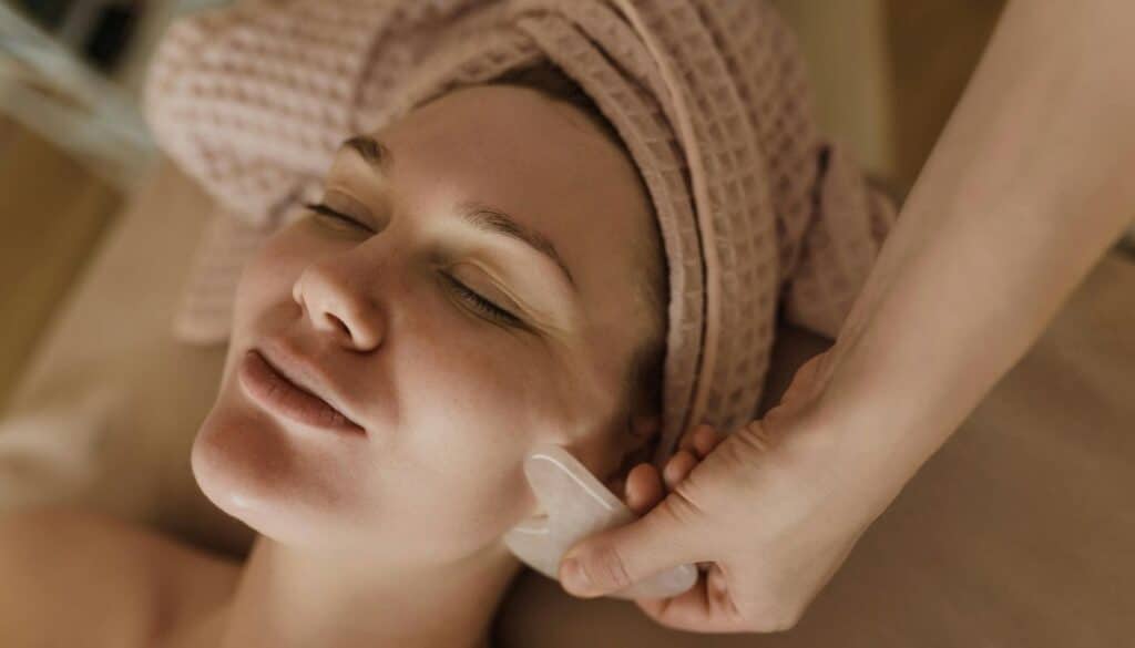 Client relaxes during a spa facial treatment while lying on a treatment bed with a towel wrapped around her hair. An esthetician uses a facial massage tool along the client's cheek in a calm, softly lit spa setting.