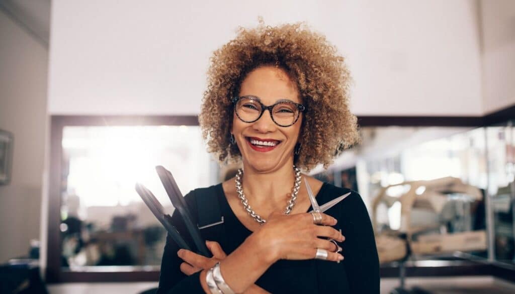 Smiling hairstylist stands in a salon holding a flat iron and haircutting shears. Wearing glasses and professional styling attire, she poses confidently in front of salon mirrors and workstations, showcasing the tools of her trade.