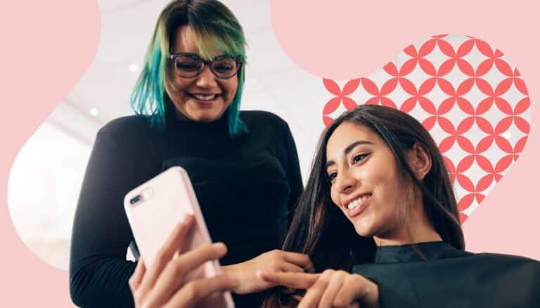 Salon professional and client smile while looking at a smartphone together during a salon appointment. The image features soft pink branded graphics and a patterned accent in the background, highlighting client engagement, personalized service, and the salon experience.