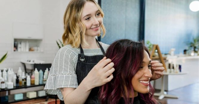 Hairstylist styles a client’s freshly colored hair while both smile in a bright, modern salon. Hair care products, plants, and salon stations are visible in the background, creating a professional and welcoming atmosphere.