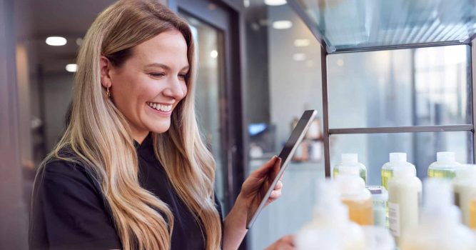 Female Stylist Or Business Owner In Hairdressing Salon Using Digital Tablet To Check Beauty Products