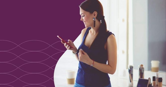 Woman stands in a salon space checking her smartphone while smiling. Beauty products, makeup brushes, and a coffee cup sit on a nearby counter, while a purple branded graphic with a subtle wave pattern frames the image, highlighting salon technology, client communication, and business growth tools.