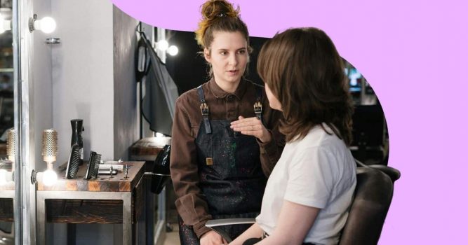 Hairstylist consults with a client seated in a salon chair, discussing hair goals and service options before an appointment. The image features a lavender branded background and a professional salon setting with illuminated mirrors, styling tools, and workstations, emphasizing personalized client consultations and salon expertise.